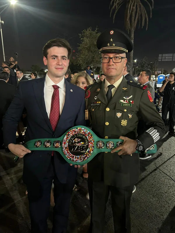 Dos asistentes, uno con uniforme militar, sostienen un cinturón WBC